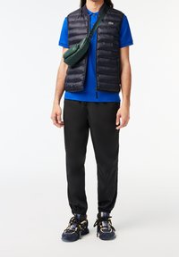 Černá prošívaná vesta přes modré polo tričko, kombinovaná s černými tepláky a vícebarevnými teniskami. Nositelná zelená taška přes rameno.