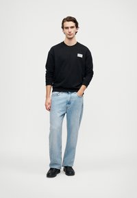 Felpa nera con collo rotondo e logo frontale, abbinata a jeans in denim azzurro chiaro. Il modello si trova con le mani in tasca, indossando scarpe nere.