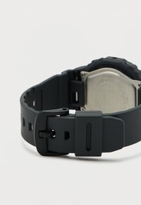 Μαύρο ψηφιακό ρολόι με μαύρο ρυθμιζόμενο λουράκι αγκράφας και εμφανές μεταλλικό πίσω κάλυμμα, τοποθετημένο σε φωτεινή επιφάνεια.