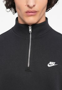 Κοντινό πλάνο ενός ατόμου που φοράει μαύρο φερμουάρ Nike με ασημί φερμουάρ που είναι μισά κλειστό και λευκό λογότυπο Nike στο στήθος.