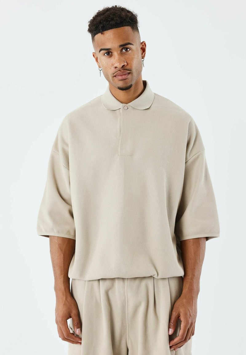 Polo beige con vestibilità rilassata, maniche corte e un singolo bottone al colletto, abbinato a pantaloni a vestibilità rilassata dello stesso colore.