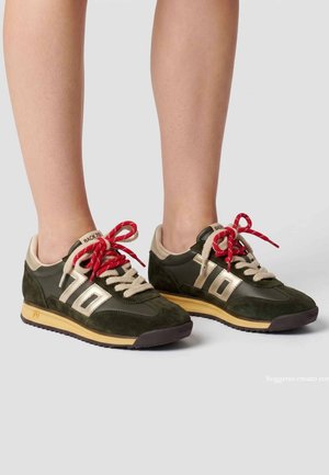 Persona che indossa sneakers verde scuro e beige con lacci rossi e beige, in piedi su una superficie grigio chiaro.