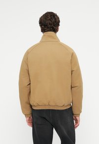 Καφέ βαμβακερό bomber τζάκετ με ψηλό γιακά, ελαστικούς καρπούς και μέση. Διαθέτει απλό σχεδιασμό χωρίς διακριτά μοτίβα ή αξεσουάρ.