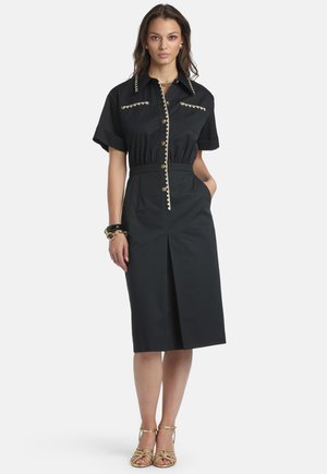 Mujer vestida con un vestido negro hasta la rodilla, de manga corta, con detalles de botones dorados, adornos en el borde y sandalias doradas de tacón alto con tiras.