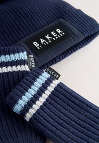 Sötétkék kötött sapka bordázott textúrával és fekete címkével. A hozzáillő borda mintás kötött kesztyűk kék-fehér csíkosak, szintén fekete címkékkel.