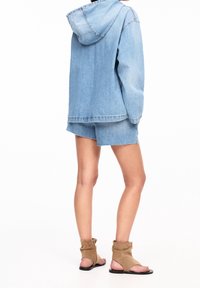 Giacca di denim azzurro chiaro con cappuccio, abbinata a pantaloni corti blu coordinati. Sandali alla caviglia in suede marrone con punte scoperte e suole nere.