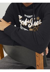 Felpa nera con testo bianco e dorato, texture morbida e cappuccio con cordino. Design elegante con polsini a coste, indossata mentre si è seduti su una sedia.