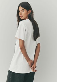 Hvid bomulds t-shirt med korte ærmer, rund halsudskæring og løs pasform, båret sammen med mørke denim bukser. Synlig tatovering på håndleddet.
