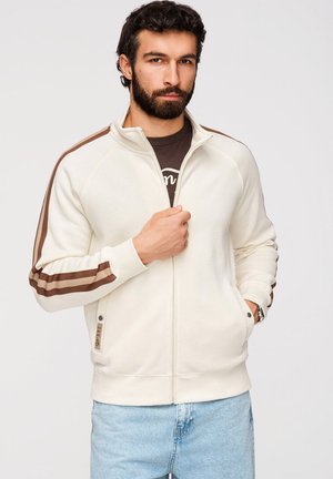 Homme aux cheveux foncés et à la barbe, portant une veste blanche zippée avec des rayures marron sur les manches et un jean bleu clair, debout devant un fond uni.