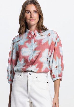 Vrouw draagt een losse, bloemrijke blouse met knopen in roze, blauw en wit met driekwart mouwen, gecombineerd met een witte broek met hoge taille, tegen een witte achtergrond.