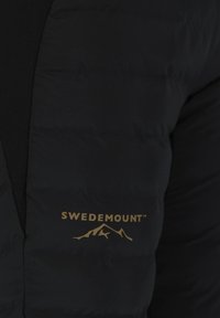Zwarte geïsoleerde jas met een gewatteerde textuur en een gladde afwerking. Bevat een gouden logo met een bergontwerp op de onderste voorkant.