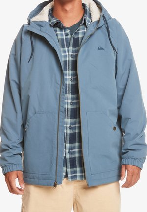 Muž v modré kapuci s fleecovou podšívkou přes modrou károvanou košili a béžové kalhoty, stojící s uvolněnými pažemi.