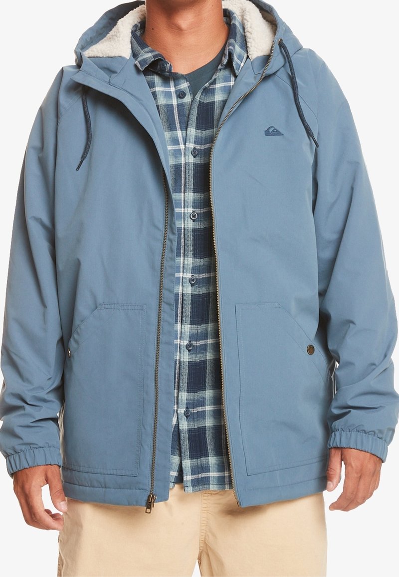 Homme portant une veste à capuche bleue avec doublure en polaire, sur une chemise à carreaux bleue et un pantalon beige, debout les bras relâchés.