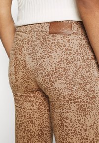 Jeans elásticos de color beige con estampado de leopardo y un tejido texturizado, con una etiqueta de cinturón de cuero marrón y dos bolsillos traseros.