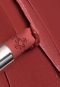 Bottiglia di rossetto opaco in rosa chiaro con logo in rilievo, che applica una texture cremosa di rosso intenso, posizionata su uno sfondo rosso ricco.