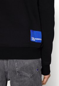 Felpa nera con polsini a costine; presenta un'etichetta blu con la scritta "KARL LAGERFELD JEANS". Abbinata a jeans grigi che mostrano il design della tasca posteriore.