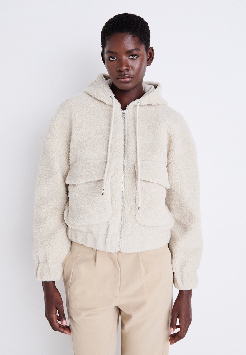 Veste en polaire de couleur crème, coupée, avec capuche, fermeture éclair à l'avant et deux grandes poches poitrine. Elle a une texture douce et des poignets élastiqués.