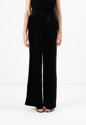 Vrouw in hoge zwarte fluwelen wijde broek met bijpassende riem en zwarte sandalen met open tenen op een witte achtergrond.