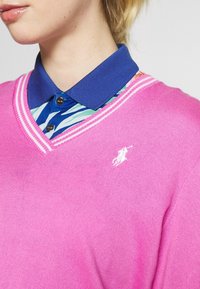 Růžový svetr s výstřihem do V a žebrovaným lemem, přes modrou košili s límečkem, která má barevný vzor listů. Malé bílé logo na svetru.