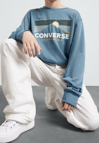 Person kneeling wearing white pants, white sneakers, and a blue sweatshirt with a mountain graphic and "Converse All Star Brand" text.