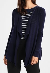 Cárdigan de punto color azul marino con un diseño de frente abierto, mangas largas y textura acanalada, superpuesto a una blusa a rayas en azul marino y blanco.