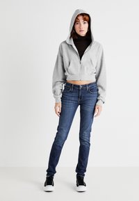 Sivý skrátený hoodie s predným zipsom a kapucňou, kombinovaný s tmavomodrými úzkymi džínsami a čiernymi vysokými teniskami. Hladké pozadie.
