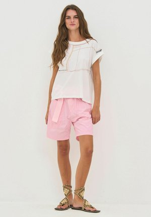 Kvinde med langt brunt hår iført en hvid top med korte ærmer, lyserøde shorts med bælte og guldnitterede sandaler, stående foran en hvid baggrund.