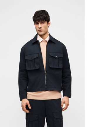 Jeune homme portant une veste noire à fermeture éclair avec de grandes poches avant, une chemise rose clair en dessous, et un pantalon cargo noir assorti.