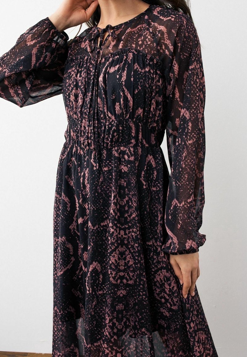 Femme portant une robe noire à manches longues en tissu transparent avec un motif abstrait rose, avec une taille froncée et un détail noué au niveau de l'encolure.