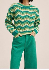 Vrouw draagt een groen, crème en tealkleurig zigzag gebreide trui met teal hoge taille wijd uitlopende broek, handen in de zakken gestoken.