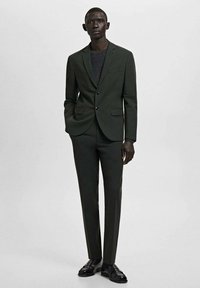 Dunkelgrüner Anzug mit figurbetontem Schnitt, einreihiger Jacke, zwei Taschen und passender Hose. Dazu ein dunkelgraues Hemd. Schwarze Schuhe.