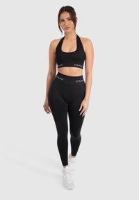 Zwarte halter crop top en high-waist leggings gemaakt van rekbare stof, met witte "SMIL" branding op de band. Model draagt witte sneakers.