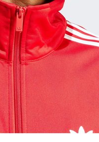 Punainen vetoketjullinen takki korkealla kauluksella, jossa valkoiset kolmiraitakuviot olkapäillä ja valkoinen logo rinnassa. Sileä kangaspinta.