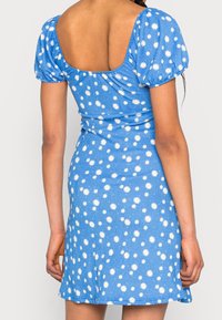Niebieska krótka sukienka z białym nadrukiem w stokrotki, bufiaste rękawy, kwadratowy dekolt z tyłu i fakturowana tkanina. Łagodnie opada nad kolano.
