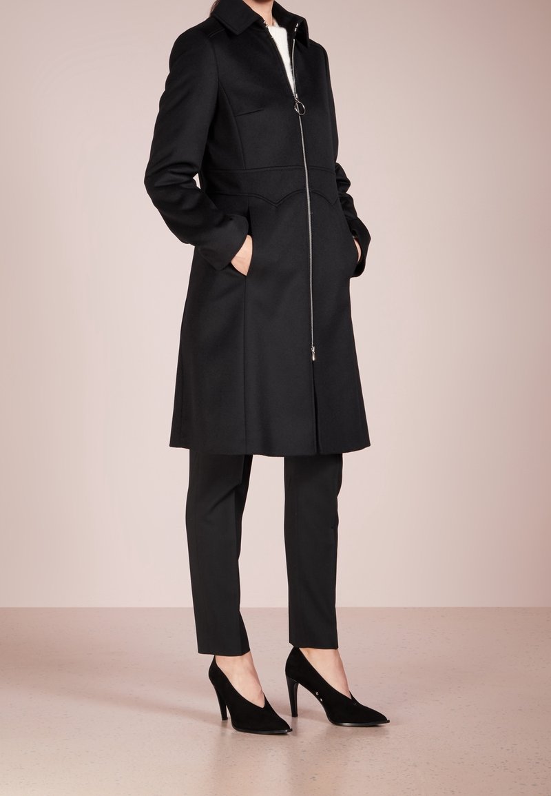 Manteau noir à la coupe structurée, fermeture éclair à l'avant, deux poches et col ; associé à un pantalon noir tailleur et des escarpins pointus.