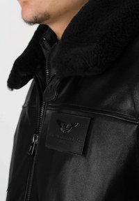 Veste en cuir noir avec un col en shearling, fermeture éclair et un patch logo texturé sur la poitrine. Fini lisse avec des coutures robustes.