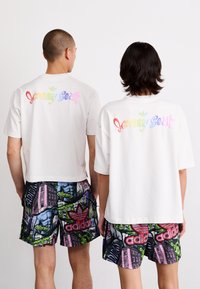 Hvide kortærmede t-shirts med regnbuefarvet 'Jeremy Scott' tekst på bagsiden. Kombineret med farverige mønstrede shorts med grafik og branding.