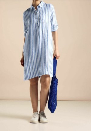 Hellblau-weiß gestreiftes Leinenhemdkleid mit Kragen, hochgekrempelten Ärmeln und einer Vordertasche, kombiniert mit einer blauen gehäkelten Tasche und weißen Schuhen.