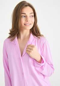 Rosa pyjamaströja med knappknäppning, krage, ficka och vit kantning längs kanterna. Tillverkad av mjukt, lätt tyg.
