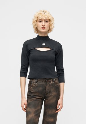 Personne blonde portant un haut court noir avec une découpe sur la poitrine et un col montant, assorti à un pantalon délavé noir et marron.