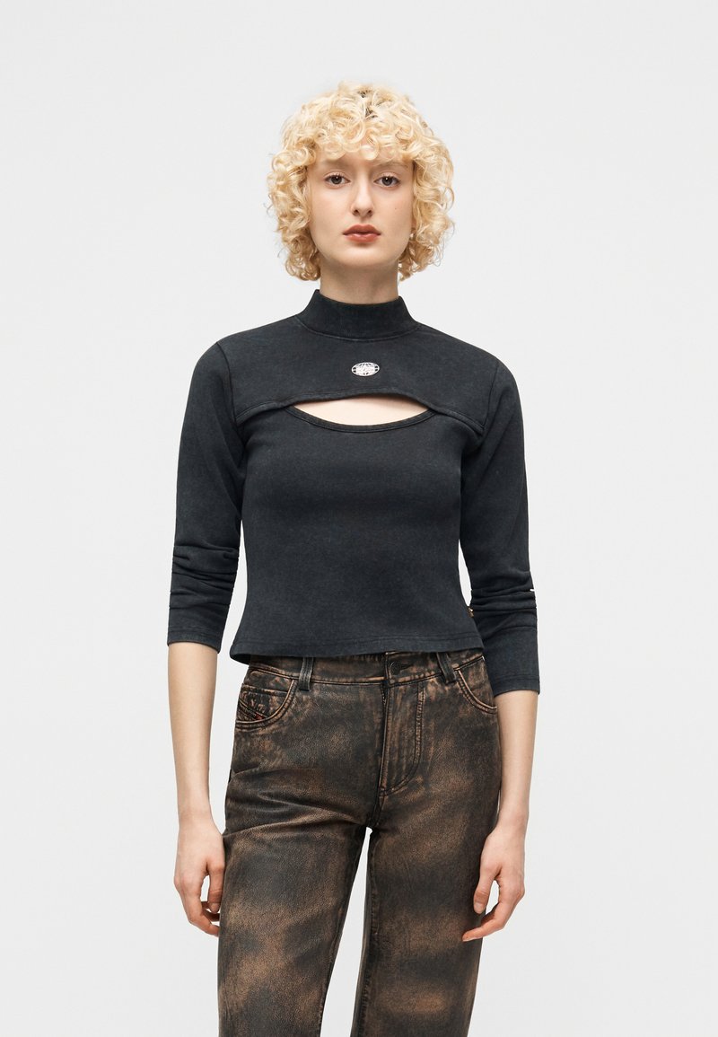 Ξανθός/ή που φοράει μαύρο κοντό τοπ με άνοιγμα στο στήθος και ψηλό λαιμό, συνδυασμένο με ξεθωριασμένο μαύρο και καφέ παντελόνι.