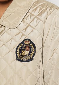 Béžová prošívaná bunda s námořnickou a zlatou vyšívanou nášivkou znaku Ralph Lauren na hrudi poblíž límce.