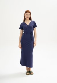 Navyblaues Wickelkleid aus weichem Stoff mit kurzen Ärmeln, einem gebundenen Taillenbereich und einem bodenlangen Schnitt. Getragen mit grünen Rutschsandalen.