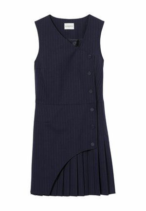 Joven mujer con cabello largo y oscuro que lleva una blusa azul a rayas con un lazo y un vestido plisado asimétrico azul marino con zapatos marrones y calcetines rojos.