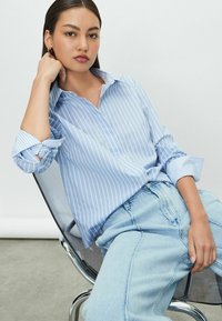 Chemise à rayures bleues et blanches avec col, manches retroussées et coupe décontractée, associée à un jean en denim bleu clair. Design moderne, texture lisse.
