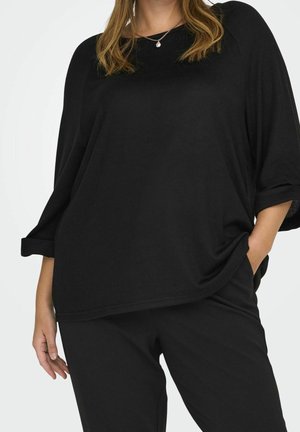 Mujer con blusa negra de manga larga y holgada, pantalones negros y mano en el bolsillo, lleva un collar dorado con un pequeño colgante.