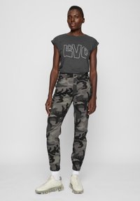 Pilkas marškinėliai su "LOVE" grafika, derinami su kamufliažo rašto siauro kirpimo kelnėmis. Modelis apsimovęs lengvus sportbačius su permatomomis padais ir baltas kojines.