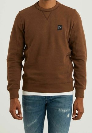 Mężczyzna w brązowej bluzie z okrągłym dekoltem, z małą czarną naszywką z logo na piersi, nałożonej na białą koszulę, w połączeniu z niebieskimi dżinsami.