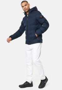 Marinblå puffjacka med huva, med dragkedjestängning och sidofickor, kombinerad med vita jeans och svarta sneakers. Slät textur.
