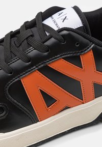 Svart lädersneaker med stort orange 'AX'-logotyp, svarta snören och texturerad gummisula med en beige accent i botten.
